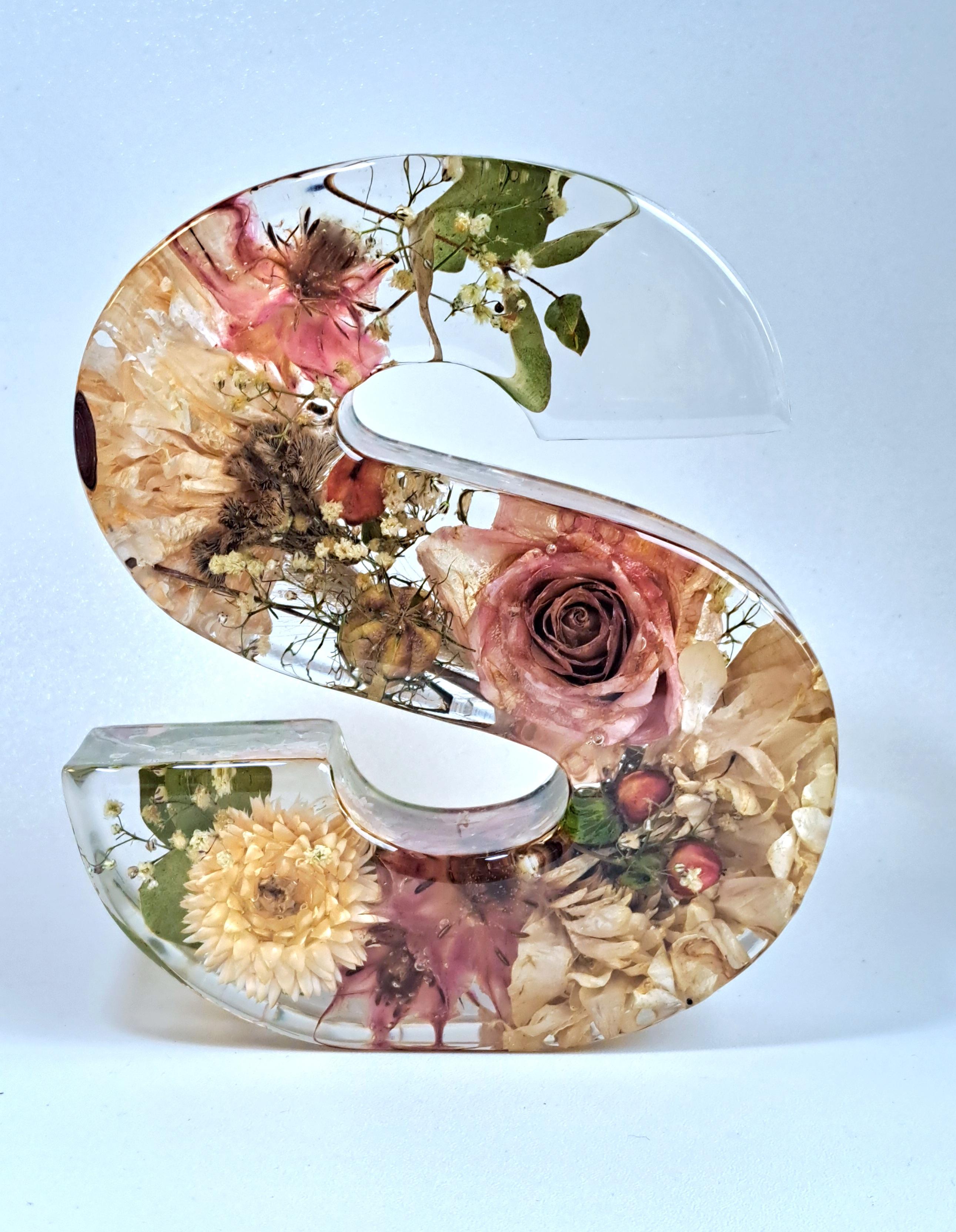 Buchstabe S aus klarem Epoxidharz mit eingegossenen Blüten. Untergrund und Hintergrund einheitlich in bläulichem weiß.