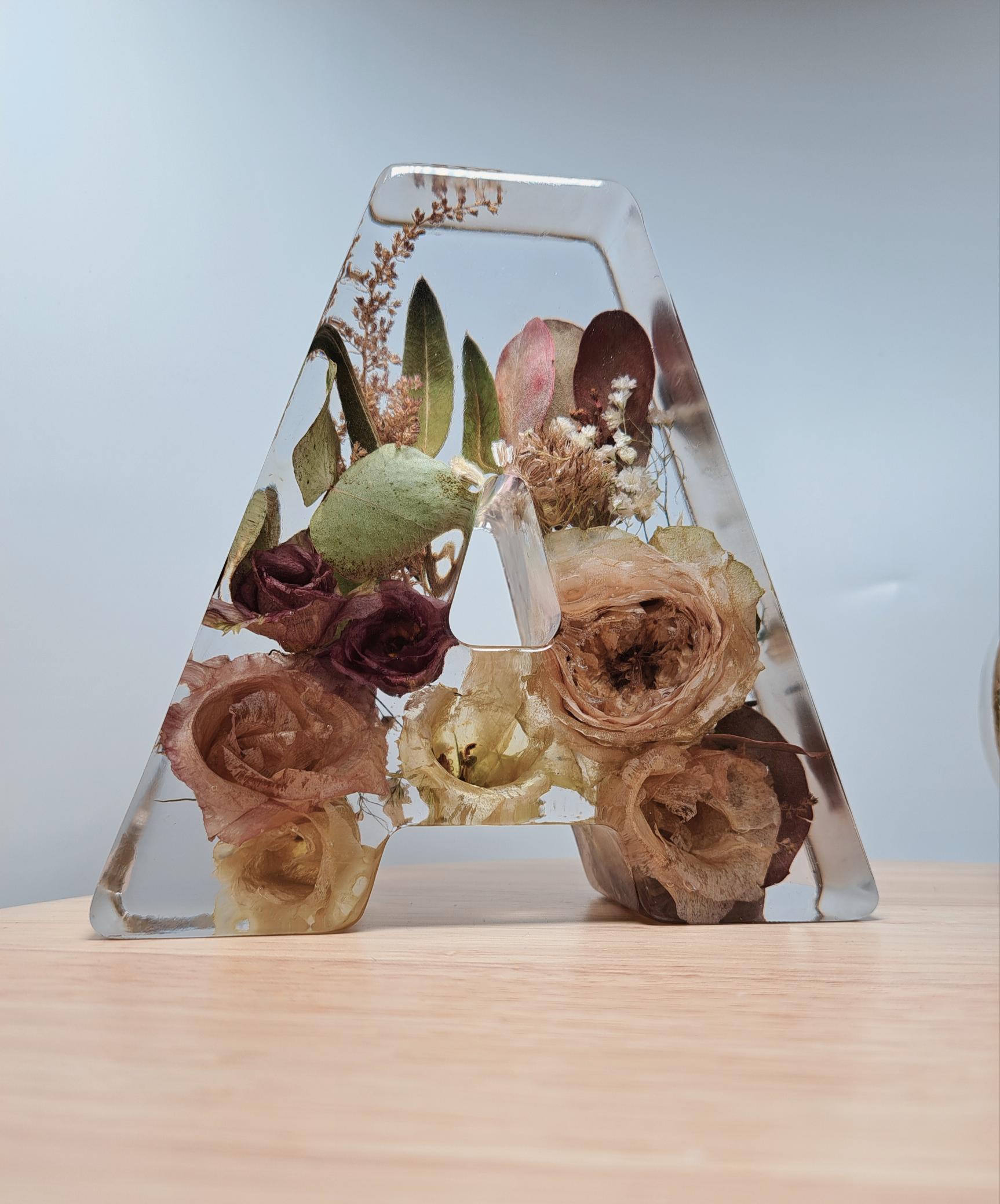 Das A steht auf einem Untergrund aus hellem Holz und vor einem weiß-gräulichen Hintergrund. Die Blumen sind rund um den Ausschnitt in der Mitte platziert und sehr gut einzeln zu sehen.