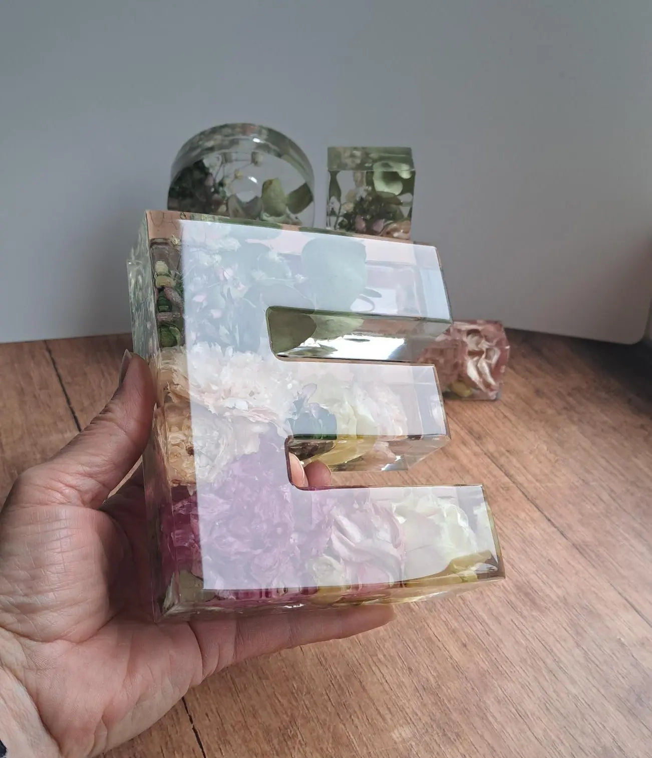 Ein in der Hand gehaltenes, 15 cm hohes E aus Epoxidharz und getrockneten Blumen aus dem eigenen Brautstrauß. Im Hintergrund sind ein &-Zeichen und ein L im gleichen Design zu erkennen. Das  ist nach rechts geneigt, weil von dort das Licht einfällt und die Oberfläche somit schön reflektiert.