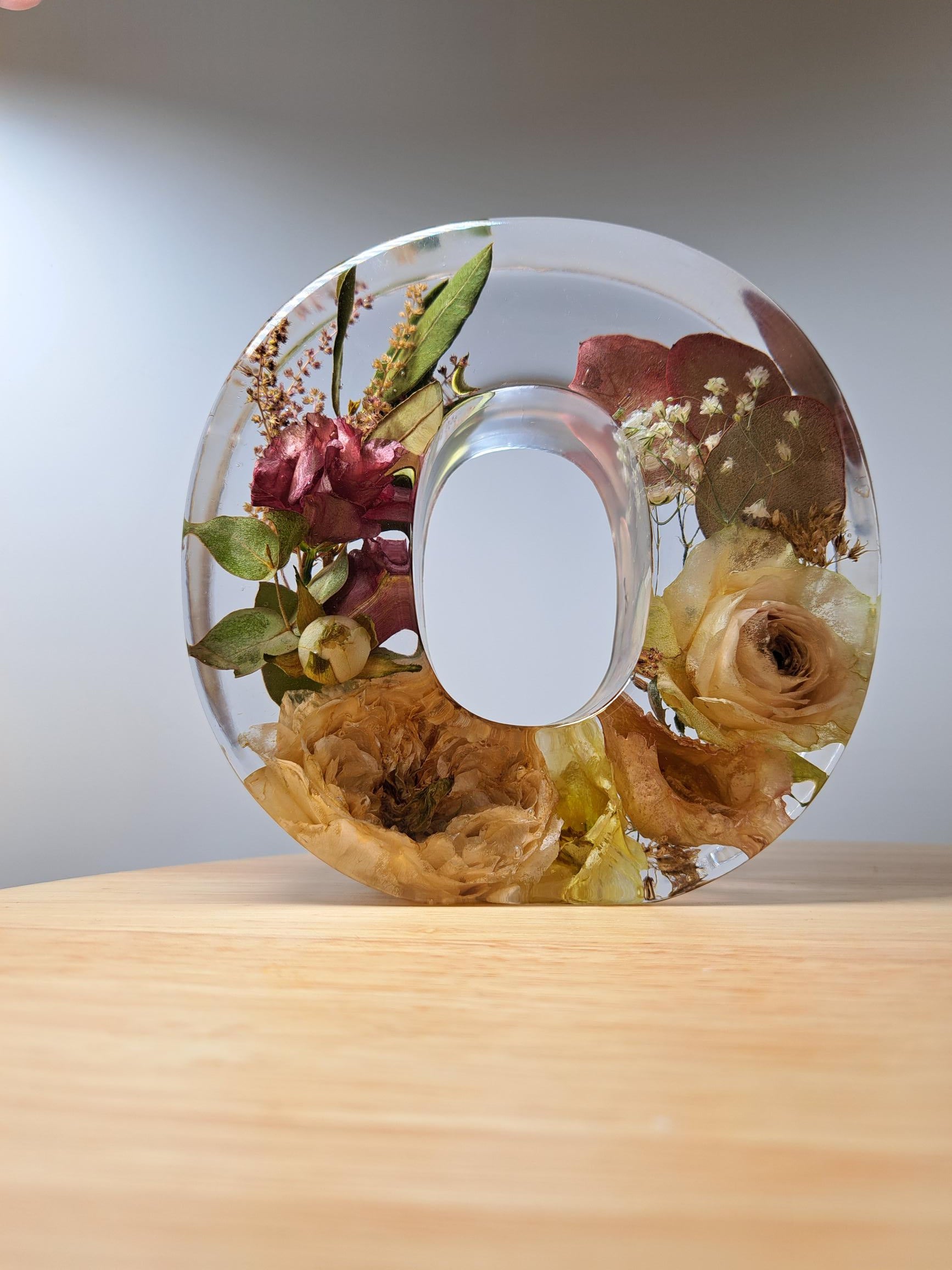 Hochwertig verarbeitetes O aus Epoxidharz mit eingearbeiteten, getrockneten Blumen aus dem eigenen Brautstrauß. Auf Holzuntergrund und weißem Hintergrund. Lichteinfall von links oben. 