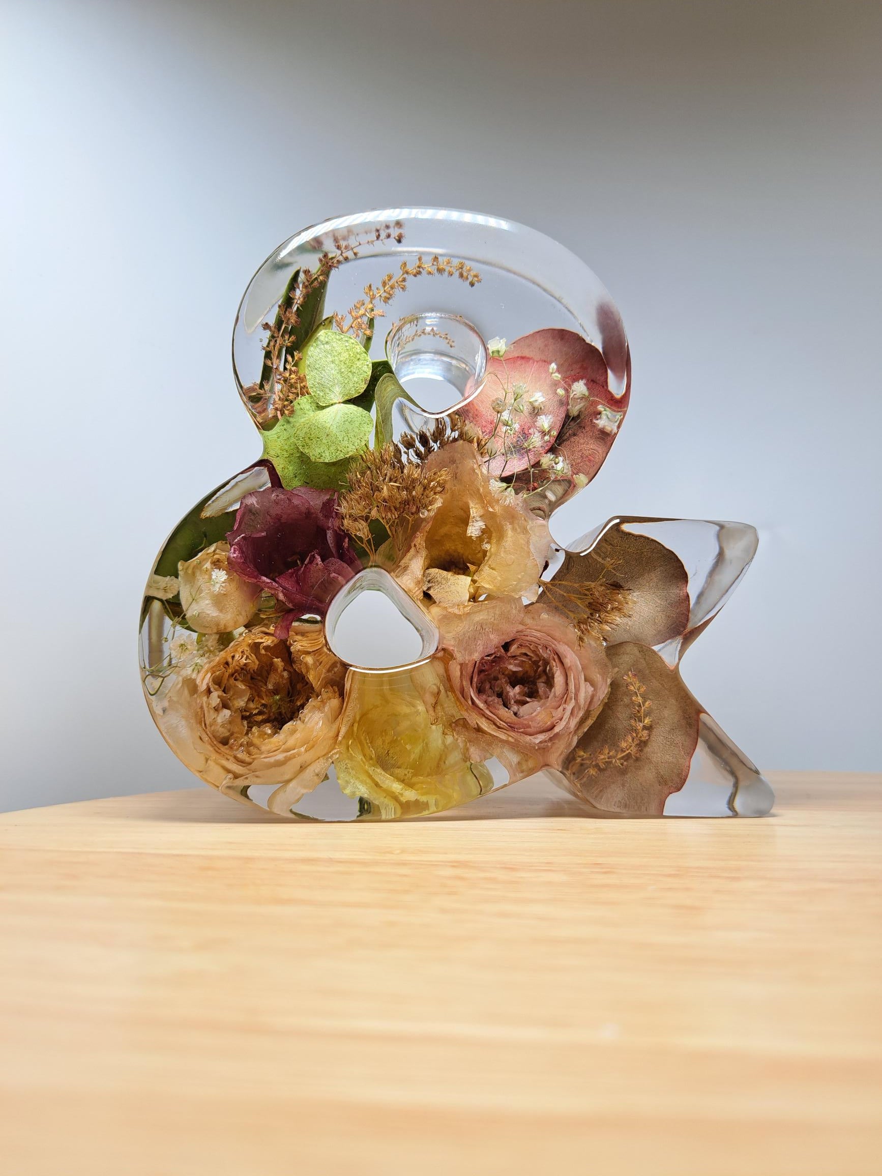 &-Zeichen aus glasklarem Epoxidharz mit eingearbeiteten Blumen aus dem eigenen Brautstrauß. Lichteinfall von links oben. Dadurch sehr schöne Beleuchtung der Blumen im Harz.